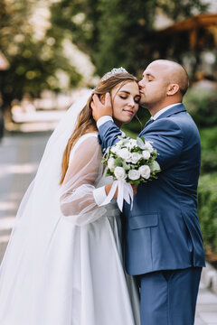 An adult stylish groom in a blue suit and a beautiful smiling bride in a white dress are hugging on a city street in a park. Wedding photography, portrait.