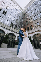 A stylish groom in a blue suit and a beautiful bride in a white dress are hugging while standing in the city, in a park against the backdrop of a multi-storey building. Wedding photography, portrait.