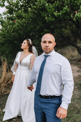 A stylish, loving groom and a beautiful, young, smiling bride in a white dress are walking in a park in nature, holding hands. wedding photography.