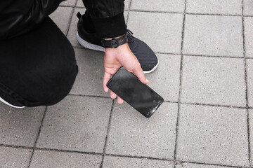 Close up view of kneeling man in jeans and shoes picking up broken phone on paved sidewalk outdoors