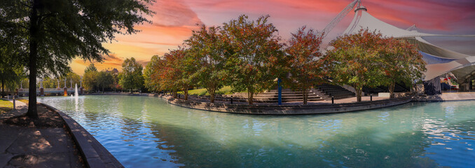 a stunning autumn landscape at World's Fair Park with a pool with blue water surrounded by red and yellow autumn trees and lush green trees and plants with a red sky at sunset in Knoxville - obrazy, fototapety, plakaty