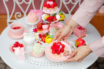 Serving festive table of sweet desserts, fresh strawberry and raspberry, tasty drinks. Person hand hold plate of delicious berry cake in cafe top view. Party in pink style, holiday, luxurious service