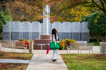 an African American woman with long sisterlocks wearing white and pink clothes, sunglasses and an orange head scarf holding and green purse walking along a footpath surrounded by autumn colored trees - obrazy, fototapety, plakaty