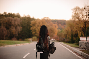 Back view of young female traveler wearing leather jacket and backpack standing on the road looking away during sunset in autumn - Powered by Adobe