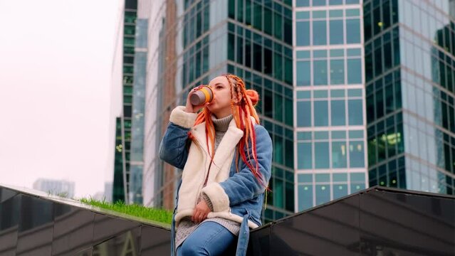 Young woman with dreadlocks drinking coffee on background of skyscrapers. Relaxed female hipster enjoying hot drink, sitting on stairway in downtown.