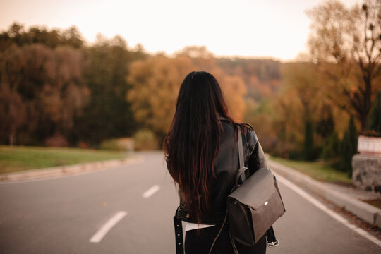 Back view of young woman wearing leather jacket and backpack walking on the road looking away during sunset in autumn