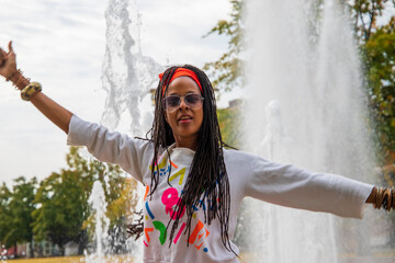 an African American woman with long sisterlocks wearing white and pink clothes, sunglasses and an orange head scarf holding dancing in front of a water fountain with lush green trees - obrazy, fototapety, plakaty