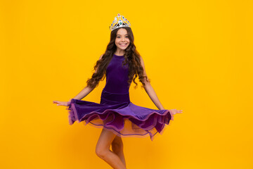 Movement windy dress. Teenage selfish girl celebrates success victory. Teen child in queen crown isolated on yellow background. Princess in tiara. Prom party. © Olena