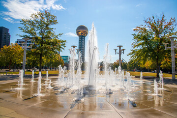 a gorgeous autumn landscape at World's Fair Park with a water fountain surrounded by autumn colored trees, lush green trees and the Sunsphere with blue sky and clouds in Knoxville Tennessee USA - obrazy, fototapety, plakaty