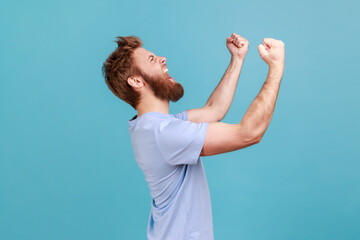Side view of overjoyed bearded man standing with excited expression, raising fists, screaming,...