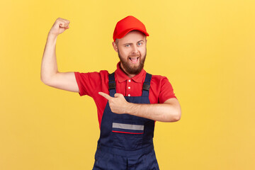 Portrait of proud strong worker man wearing blue uniform and red cap standing with raised arms and...