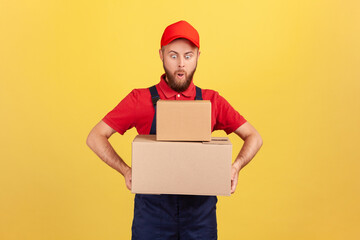 Amazed surprised deliveryman in uniform holding two heavy big cardboard boxes in hands, having funny facial expression, delivering oreder. Indoor studio shot isolated on yellow background.
