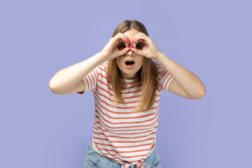 Portrait of shocked astonished blond woman wearing striped T-shirt standing and looking though binoculars, having shocked facial expression. Indoor studio shot isolated on purple background.
