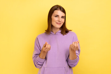 Attractive smiling young adult woman standing and looking at camera with money or italian gesture with hand, wearing purple hoodie. Indoor studio shot isolated on yellow background.