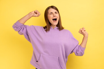 Portrait of young adult sleepless woman yawning and raising hands up, feeling fatigued, standing with close eyes, wearing purple hoodie. Indoor studio shot isolated on yellow background.