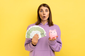 Portrait of shocked amazed woman with dark hair holding big sum of money and piggy bank, profitable investment, wearing purple hoodie. Indoor studio shot isolated on yellow background.