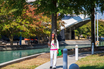 an African American woman with long sisterlocks wearing white and pink clothes, sunglasses and an orange head scarf holding an IPhone walking along a footpath near a pool surrounded by autumn trees - obrazy, fototapety, plakaty