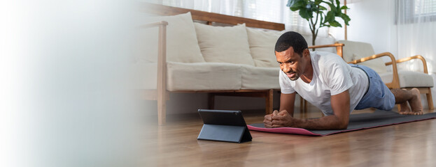 Black African American man looking at digital tablet and doing plank exercise in Living room at home. Wellness, Fitness and healthy lifestyle.