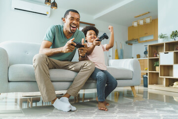 Cheerful Black African American Father and his Little son in Afro hair sitting on sofa using joysticks or game controllers playing console video games at home