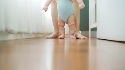 Parent teaching little baby learning to walk on wooden floor at home. Toddler enjoying the first steps with family. Chubby legs, Close up