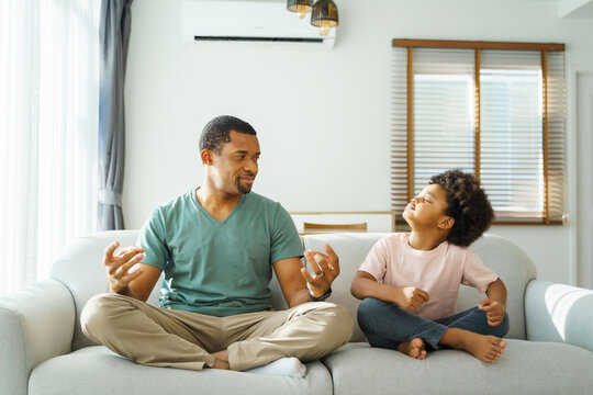 Black African american father and Playful little boy sitting on sofa doing yoga in meditation position at home together.