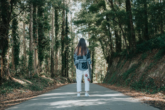 Full Body Portrait Young Woman From Behind Walking With Long Board In The Middle Of The Forest.