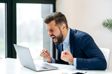 Angry annoyed frustrated caucasian business man, sitting at workplace with laptop, screaming, gesturing with fists, failed deal, disappointed with project result, experiencing negative emotions