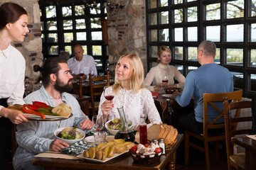 Fototapeta na wymiar Beautiful couple eating out at a restaurant while young and obliging waitress is serving them dishes. High quality photo