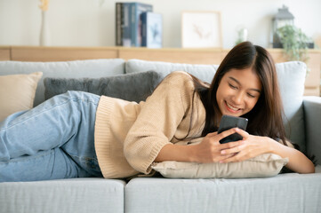 Happy young asian woman relax on comfortable couch at home texting messaging on smartphone, smiling girl use cellphone, browse wireless internet on gadget, shopping online from home