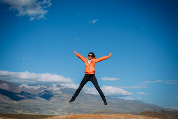 Young female traveller jumping with hands up against the mountains and blue sky. Happy woman enjoying active vacation. Travel and fun concept.