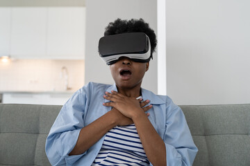 Excited African American woman wearing VR technology headset gets scaredwatching movie with futuristic 3d effect. Lifestyle of black emotional girl sits on sofa addicted to video games or metaverses 