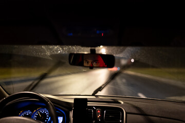 Eyes of young man are reflected in rearview mirror during night trip around city. Taxi driver or...