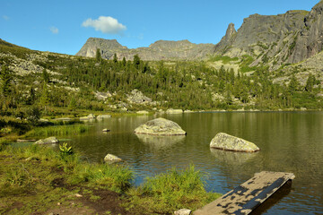 A wooden platform on the rocky shore of a picturesque lake surrounded by beautiful mountains with fragments of a coniferous forest.