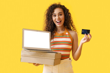 Happy young African-American woman with credit card, parcel and laptop on yellow background. Online...