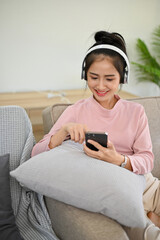 Obraz premium Pretty Asian female listening to music through headphones while relaxing in living room.