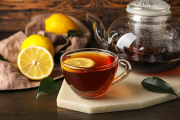 Board with glass cup of black tea, lemon and teapot on dark wooden background, closeup
