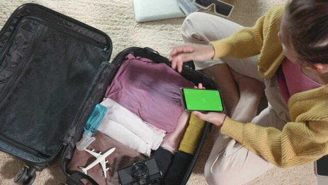 Woman using smartphone blank green screen for checklist and packing suitcase bag for travel trip weekend vacation at home, female check note purveyance or baggage on mobile phone