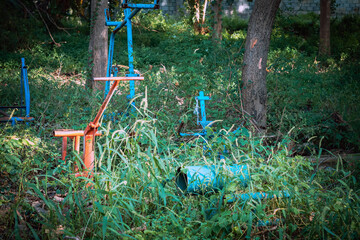 Old outdoor exercise equipment so worn out and covered in trees in outdoor exercise park. Old abandoned gym equipment on sports ground with overgrown grass. Unmaintained abandoned playground.