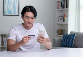 Fototapeta premium Young Asian man holding credit card and using smartphone. Businessman working at home. Online shopping, e-commerce, internet banking, spending money, working from home concept