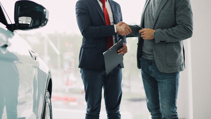 Motor showroom employee is giving car key to young male buyer and shaking hands, dealer is holding documents and gesturing. Selling and buying luxury transport concept.