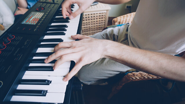 Young bearded keyboardist is playing keyboard while his friends during rehearsal at home. Close shot of hands on key buttons.