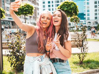 Two young beautiful smiling hipster female in trendy summer clothes.Sexy carefree women posing in the street with pink hair. Positive pure models having fun at sunset. Cheerful and happy. Take selfie