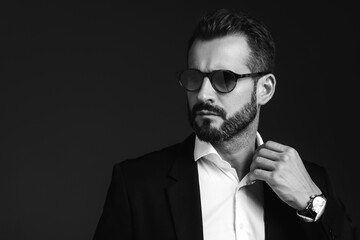 Fototapeta premium Closeup portrait of handsome confident stylish hipster lambersexual model. Sexy modern man dressed in elegant black suit. Fashion male posing in studio on dark background. In sunglasses