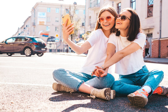 Portrait of two young beautiful smiling hipster female in trendy summer white t-shirt clothes.Sexy carefree women posing on street background. Positive models having fun, taking selfie photos