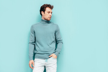 Portrait of handsome confident  model. Sexy stylish man dressed in  sweater and jeans. Fashion hipster male with curly hairstyle posing near blue wall in studio. Isolated