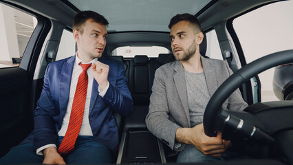 Young confident businessman is discussing new car with professional salesman sitting in front seats...