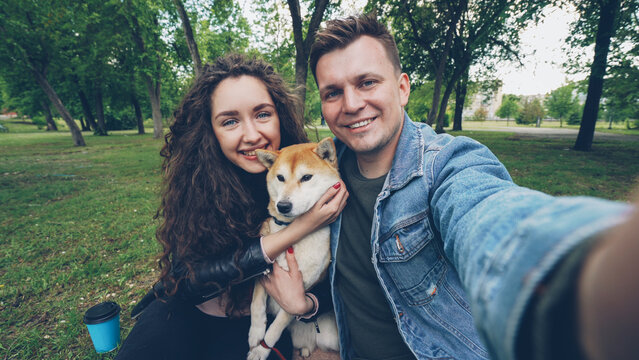 Young wife and husband are taking selfie with adorable dog and hugging each other and the animal. Point of view shot of happy people, pet and green park.