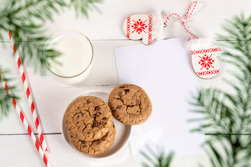 Top view through green fir branches to milk glass, straws, cookies, empty page, mitts on white table