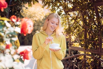 Blonde happy woman smile and drinks coffee at sweater weather warm autumn day. Blured red roses and fall nature park background. Dreamy girl holding cup in hands.