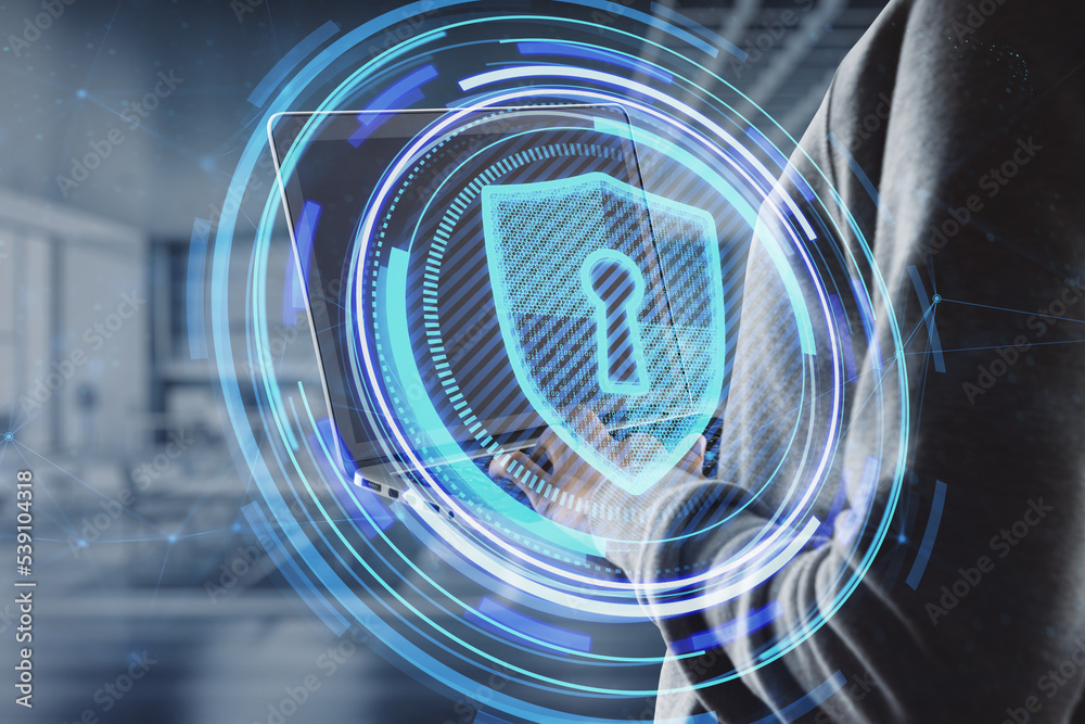 Poster close up of hacker hand using laptop with glowing shield hologram on blurry office interior backgrou